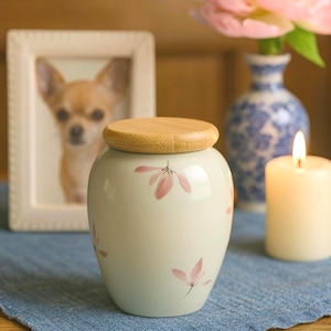 May include: A small, off-white ceramic urn with a wooden lid, adorned with pink floral designs. A framed photo of a dog is in the background, along with a lit candle and a vase with flowers. The urn is sitting on a blue cloth.