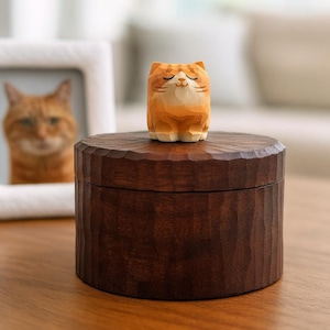 Peut inclure: Une petite figurine de chat tigré orange sculptée à la main en bois, les yeux fermés, est posée sur une boîte cylindrique en bois brun foncé. La boîte présente une surface texturée et facettée sur les côtés et le couvercle.