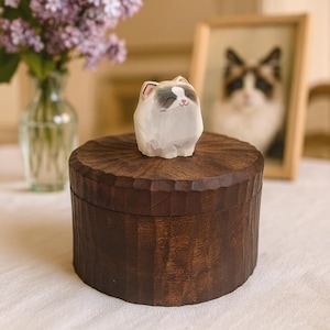 Pode incluir: Uma caixa cilíndrica de madeira castanho-escura com uma superfície esculpida e texturizada. Uma pequena figura de madeira esculpida à mão de um gato branco e cinzento com olhos fechados e nariz cor-de-rosa está fixada no centro da tampa.
