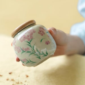 Puede incluir: Un pequeño tarro de cerámica color crema con tapa de madera. El tarro está decorado con motivos florales rosas y hojas verdes. El tarro está sostenido en la mano de una persona.