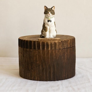 May include: A small, hand-painted figurine of a tabby cat with white chest fur and a light green collar featuring a black charm, perched atop a cylindrical, dark brown wooden box with a vertically textured, carved exterior surface.