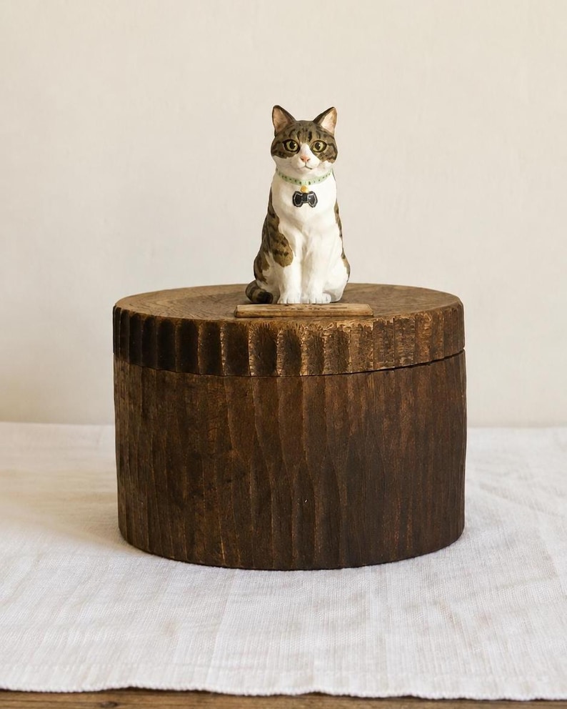 May include: A small, hand-painted figurine of a tabby cat with white chest fur and a light green collar featuring a black charm, perched atop a cylindrical, dark brown wooden box with a vertically textured, carved exterior surface.