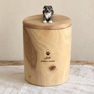 Puede incluir: Una urna cil&iacute;ndrica de madera clara con una peque&ntilde;a figura pintada a mano de un perro gris y blanco sobre la tapa. La parte frontal de la urna est&aacute; grabada con una huella, el nombre Jett y las fechas 25/5/2015 - 3/7/2026.