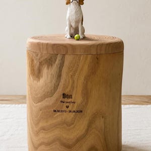 Puede incluir: Una urna cil&iacute;ndrica de madera con una figura de beagle tallada y una peque&ntilde;a pelota verde encima. El frente est&aacute; grabado con el texto "Ben", "The best boy", un s&iacute;mbolo de coraz&oacute;n y las fechas "06.02.2013 - 26.06.2026" en letras oscuras.
