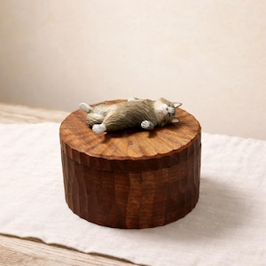 May include: A round, dark brown wooden box with a textured, carved surface. Resting on the lid is a small, detailed figurine of a tabby cat lying on its back with its eyes closed, featuring brown and grey fur with white paws and a white belly.