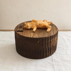 May include: A round, dark brown wooden box with a textured, scalloped side surface. Resting on the flat lid is a small, sculpted orange tabby cat lying on its side with eyes closed, positioned next to a small, rectangular dark wooden plaque.