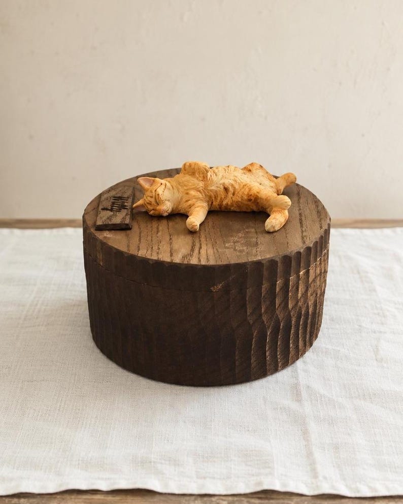 May include: A round, dark brown wooden box with a textured, scalloped side surface. Resting on the flat lid is a small, sculpted orange tabby cat lying on its side with eyes closed, positioned next to a small, rectangular dark wooden plaque.