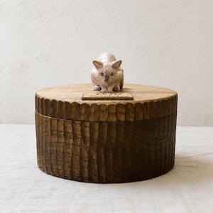 May include: A round, dark brown wooden box with a textured, vertically grooved exterior. A small, light-colored carved cat figurine sits on the lid, positioned behind a rectangular plaque engraved with the text OTTO LOVE YOU SO MUCH FOREVER.