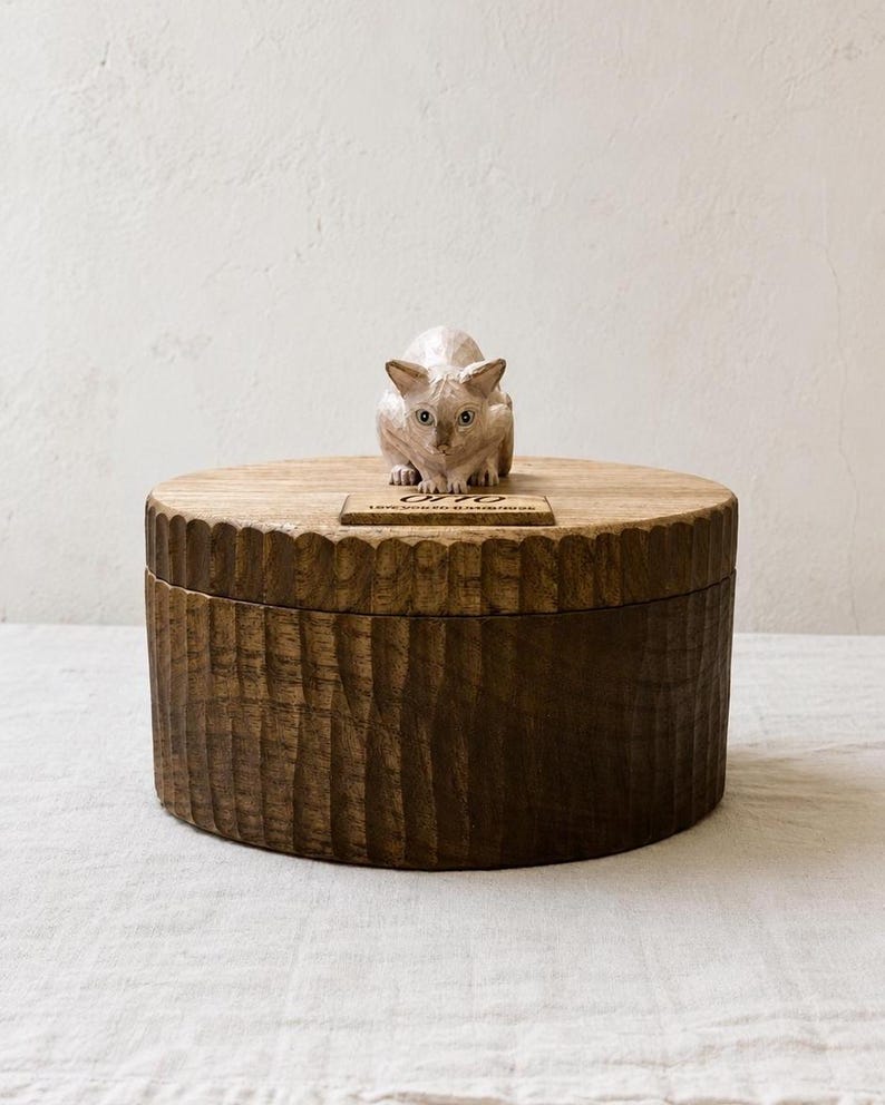 May include: A round, dark brown wooden box with a textured, vertically grooved exterior. A small, light-colored carved cat figurine sits on the lid, positioned behind a rectangular plaque engraved with the text OTTO LOVE YOU SO MUCH FOREVER.