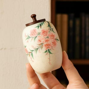 May include: A small, off-white ceramic jar with a dark brown wooden lid. The jar is decorated with pink cherry blossom flowers and green leaves. The jar has a rounded shape and is held in a person's hand.