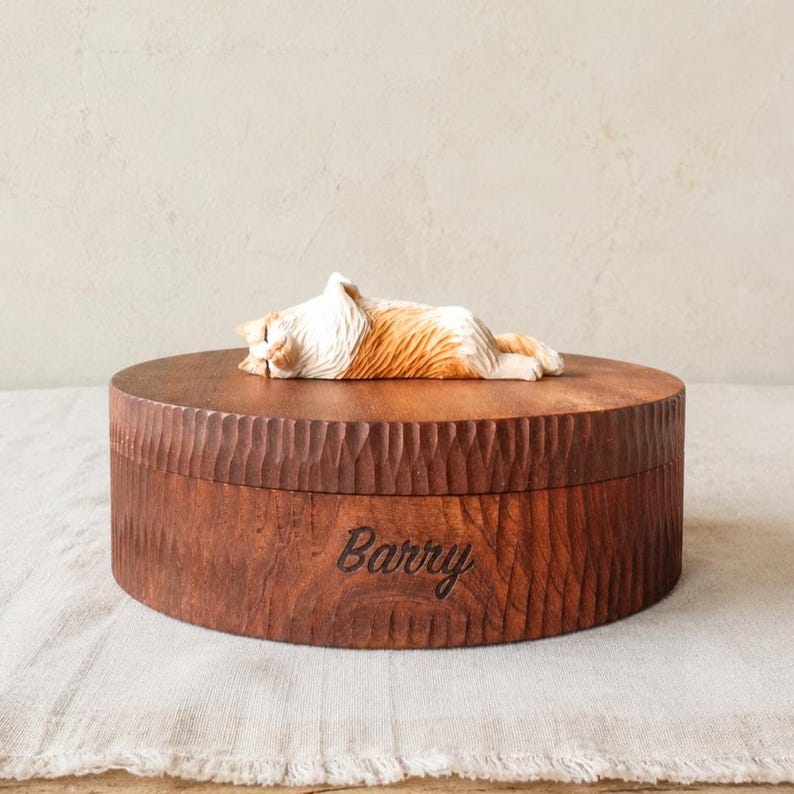 May include: A round wooden box with a textured, carved exterior and the name Barry engraved on the side. Resting on top of the lid is a small, detailed figurine of a sleeping orange and white cat with its eyes closed and paws tucked in.