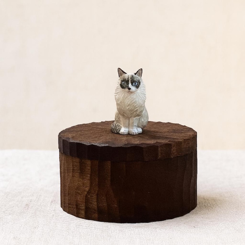 May include: A small, hand-carved wooden figurine of a cat with cream and dark brown fur, blue eyes, and a pointed face, perched atop a round, dark brown wooden box featuring a textured, scalloped edge along the lid and a vertical carved side.