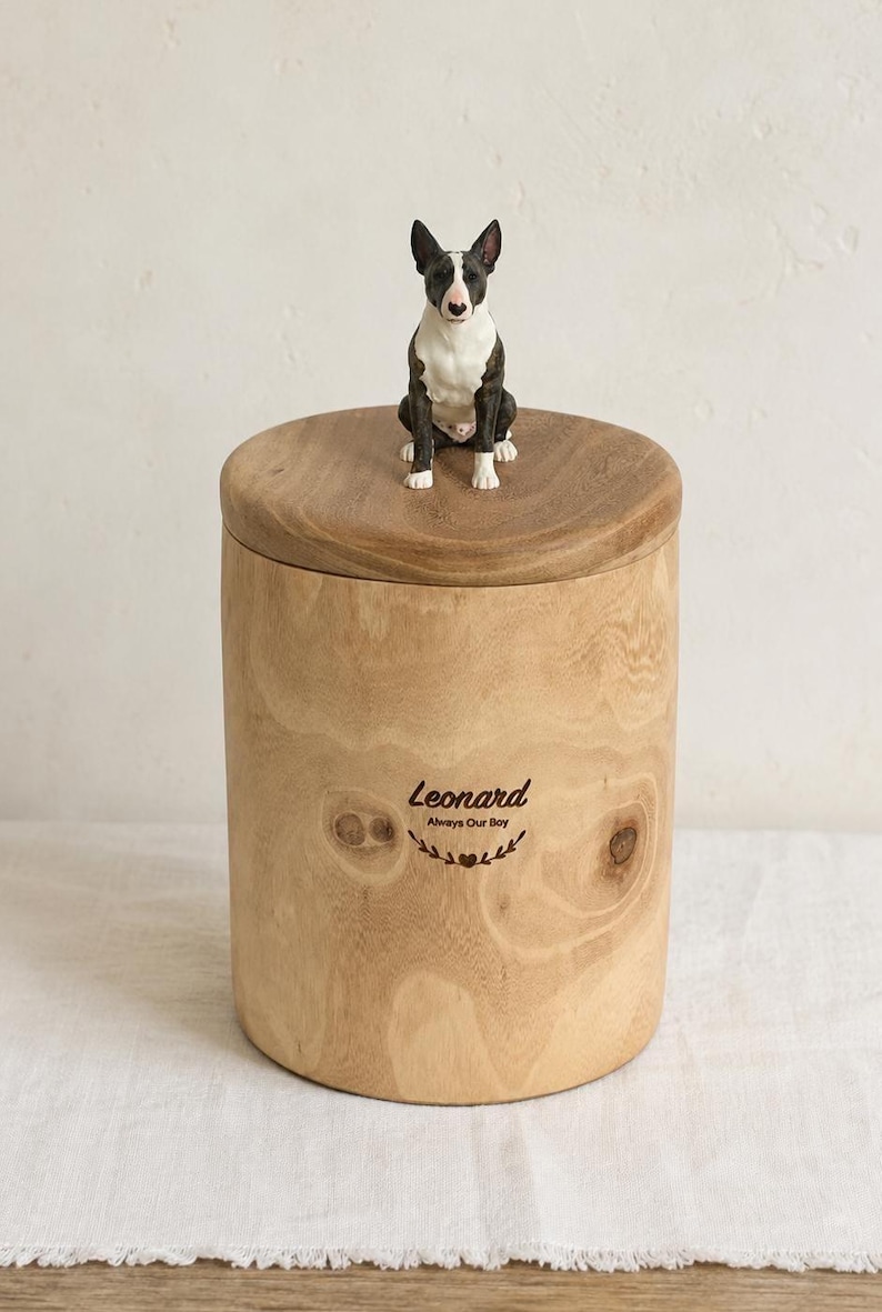 Puede incluir: Una urna cil&iacute;ndrica de madera con veta marr&oacute;n claro natural y tapa extra&iacute;ble. Una peque&ntilde;a figura pintada a mano de un Bull Terrier blanco y negro se asienta en la parte superior. El frente est&aacute; grabado con el texto Leonard Always Our Boy y un coraz&oacute;n.