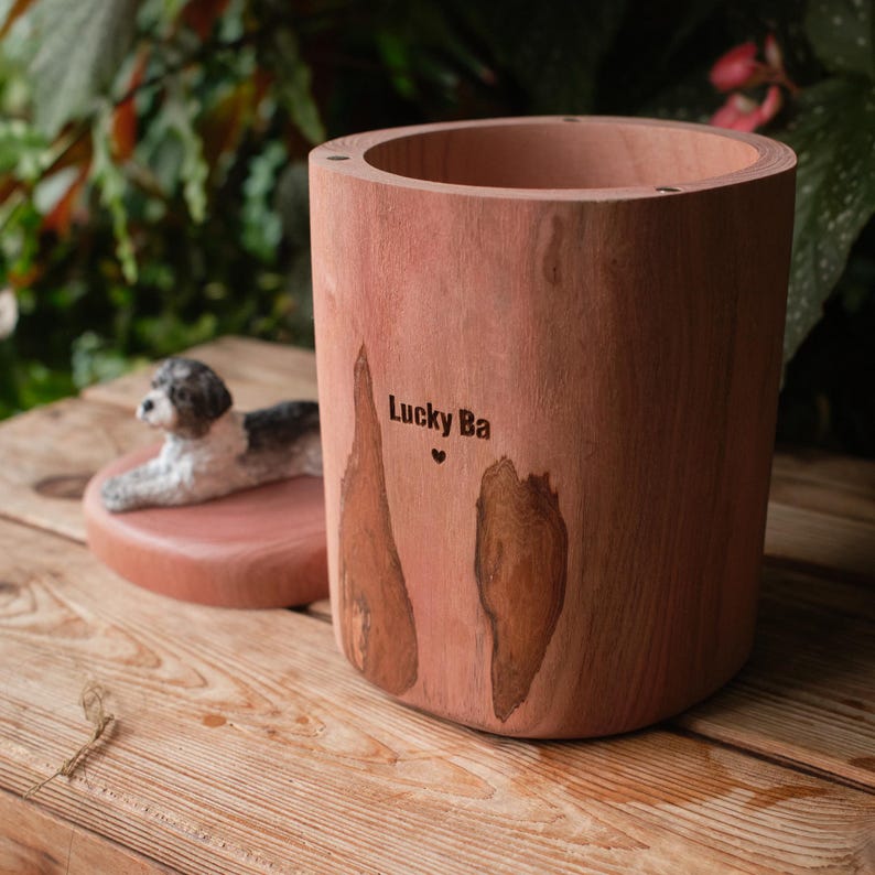 Puede incluir: Una urna cil&iacute;ndrica de madera con acabado de veta natural, que presenta el texto grabado Lucky Ba y un peque&ntilde;o s&iacute;mbolo de coraz&oacute;n. La tapa est&aacute; separada y muestra una figura esculpida y pintada de un perro blanco y negro en reposo.