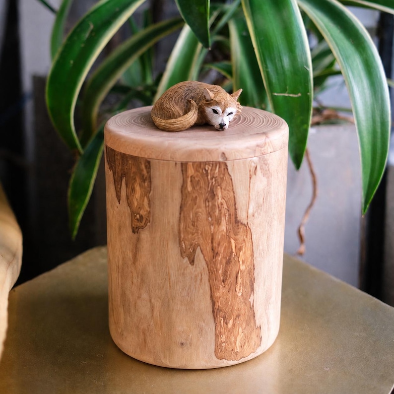 Puede incluir: Una urna cil&iacute;ndrica de madera con un acabado de veta de madera marr&oacute;n claro natural y una superficie superior lisa. Una peque&ntilde;a figura de zorro marr&oacute;n tallada a mano se encuentra en una postura acurrucada y dormida sobre la tapa del recipiente.