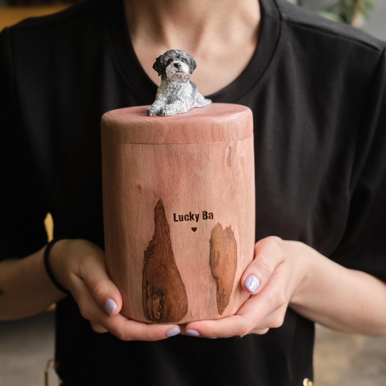 Puede incluir: Una urna cil&iacute;ndrica de madera con un acabado natural en marr&oacute;n claro y detalles de vetas de madera oscura. La tapa presenta una peque&ntilde;a figura esculpida de un perro blanco y negro. El texto "Lucky Ba" y un peque&ntilde;o coraz&oacute;n est&aacute;n grabados en el frente.