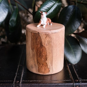 Puede incluir: Una urna cil&iacute;ndrica de madera con veta natural de color marr&oacute;n claro y acabado liso. Una peque&ntilde;a figura detallada de un perro marr&oacute;n y blanco en posici&oacute;n tumbada est&aacute; fijada en el centro de la tapa plana y circular sobre el recipiente.