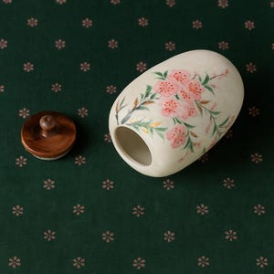 May include: A cream-colored, oval-shaped ceramic container with a wooden lid. The container is hand-painted with pink cherry blossoms and green leaves. The lid is a dark brown color and has a small wooden knob.