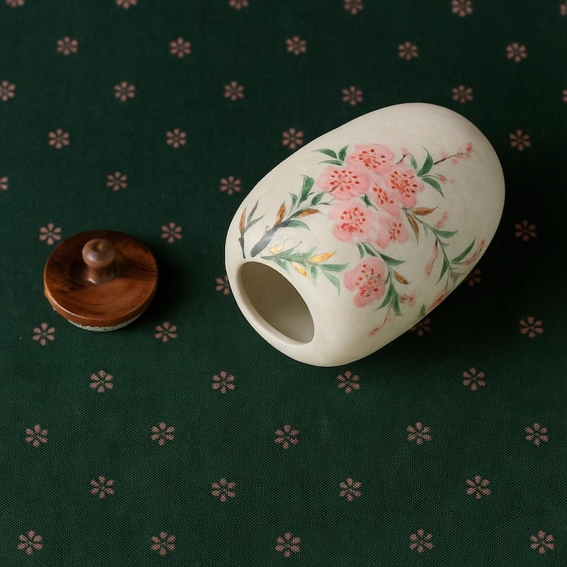 May include: A cream-colored, oval-shaped ceramic container with a wooden lid. The container is hand-painted with pink cherry blossoms and green leaves. The lid is a dark brown color and has a small wooden knob.