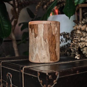 Puede incluir: Un recipiente cil&iacute;ndrico de madera con un patr&oacute;n de veta natural que presenta vetas de color marr&oacute;n claro y oscuro. El recipiente tiene una tapa superior plana y lisa y un cuerpo de lados rectos, mostrando la textura org&aacute;nica de la madera.