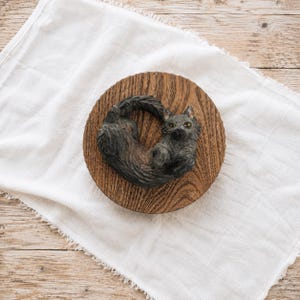 May include: A dark gray, textured cat figurine curled into a circular shape, featuring pointed ears and yellow eyes. The cat is mounted on a round, dark brown wooden base with a visible grain pattern, creating a cohesive decorative object.