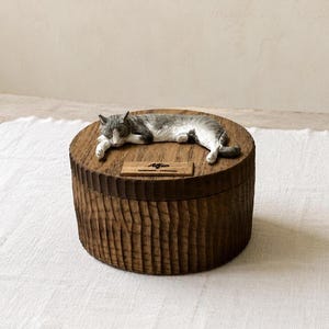 May include: A round, dark brown wooden box with a textured, vertically grooved exterior. A sculpted, grey and white cat figurine lies on the lid. A small rectangular wooden plaque on the lid features the engraved name Alfio in black script.