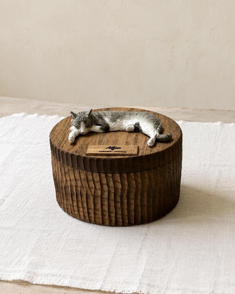 May include: A round, dark brown wooden box with a textured, vertically grooved exterior. A sculpted, grey and white cat figurine lies on the lid. A small rectangular wooden plaque on the lid features the engraved name Alfio in black script.
