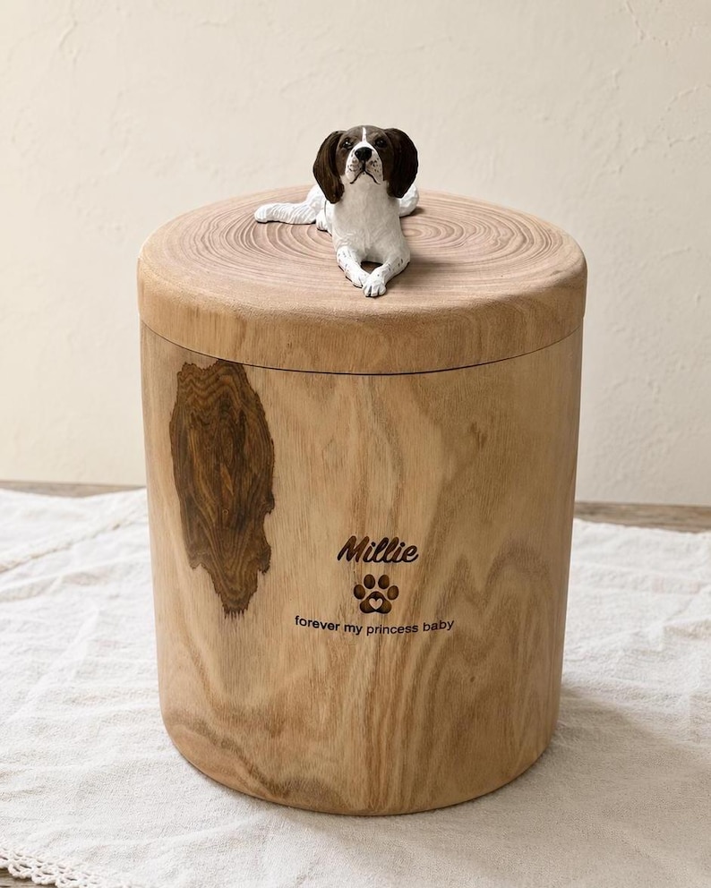 Puede incluir: Una urna cil&iacute;ndrica de madera con acabado de veta natural y un nudo oscuro en el lateral. Una figura esculpida de un perro marr&oacute;n y blanco yace sobre la tapa. El frente est&aacute; grabado con el nombre Millie, una huella y el texto forever my princess baby.