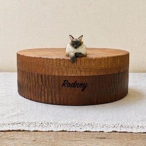 May include: A round wooden box with a textured side and a lid featuring a small, realistic Siamese cat figurine perched on top. The name Rodney is engraved in a dark script font on the side of the box, which has a warm, natural wood finish.