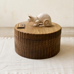 May include: A round, dark brown wooden box with a vertically fluted exterior texture. A small, light-colored, carved cat figurine sits on the lid next to a rectangular wooden plaque featuring the engraved name Otto in a script font.