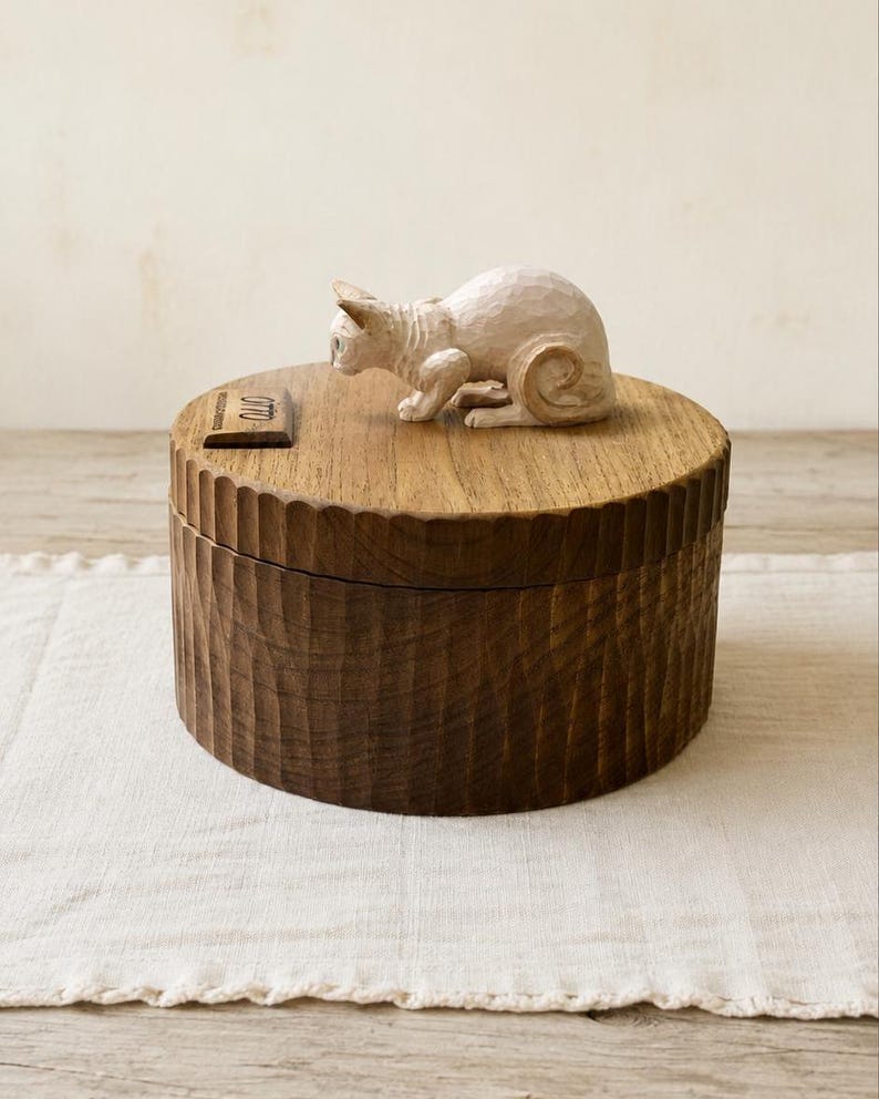 May include: A round, dark brown wooden box with a vertically fluted exterior texture. A small, light-colored, carved cat figurine sits on the lid next to a rectangular wooden plaque featuring the engraved name Otto in a script font.