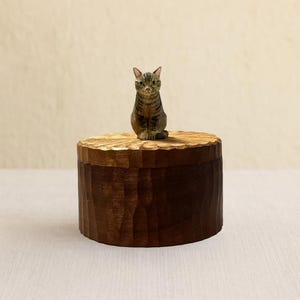 May include: A round, dark brown wooden box with a textured, carved surface. A small, hand-painted figurine of a brown tabby cat with green eyes and pointed ears is securely attached to the center of the lid, facing forward.