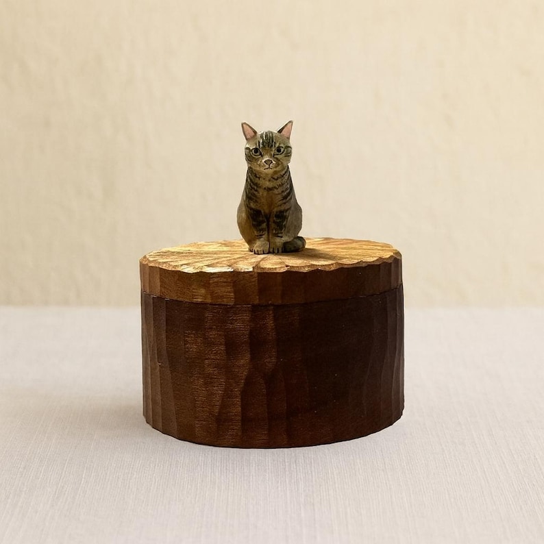 May include: A round, dark brown wooden box with a textured, carved surface. A small, hand-painted figurine of a brown tabby cat with green eyes and pointed ears is securely attached to the center of the lid, facing forward.