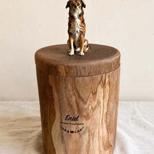 Puede incluir: Una urna cil&iacute;ndrica de madera con acabado de veta natural, rematada con una peque&ntilde;a y detallada figura de un perro marr&oacute;n y blanco. El frente de la urna presenta el nombre grabado Enid, la frase Forever in our hearts y un peque&ntilde;o icono de coraz&oacute;n.