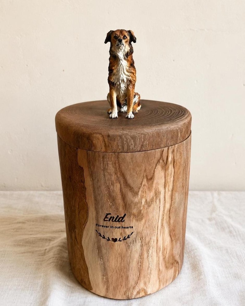 Puede incluir: Una urna cil&iacute;ndrica de madera con acabado de veta natural, rematada con una peque&ntilde;a y detallada figura de un perro marr&oacute;n y blanco. El frente de la urna presenta el nombre grabado Enid, la frase Forever in our hearts y un peque&ntilde;o icono de coraz&oacute;n.