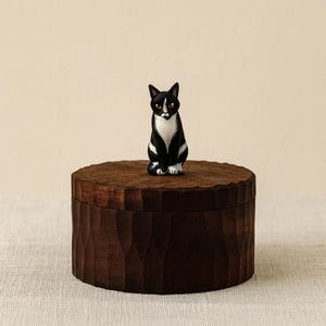 May include: A round, dark brown wooden lidded box with a textured, carved exterior surface. Perched on top of the lid is a small, hand-painted figurine of a black and white cat with yellow eyes, sitting in an upright, alert position.