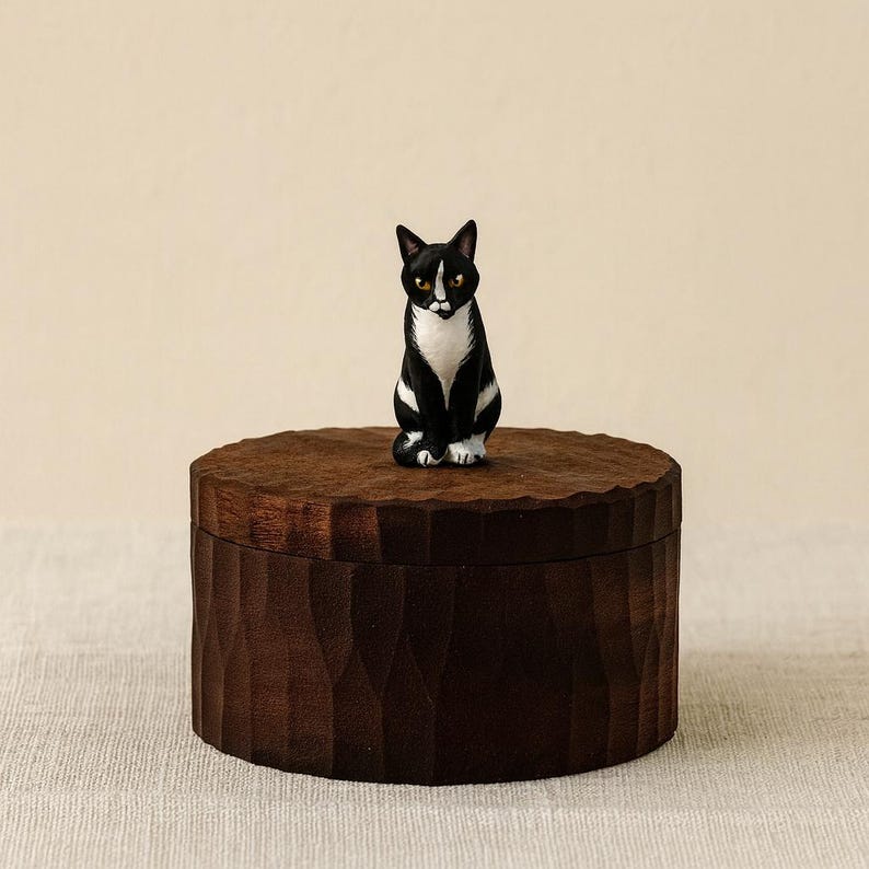 May include: A round, dark brown wooden lidded box with a textured, carved exterior surface. Perched on top of the lid is a small, hand-painted figurine of a black and white cat with yellow eyes, sitting in an upright, alert position.