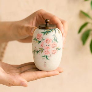 May include: A small, off-white ceramic jar with a bronze-colored lid. The jar is adorned with hand-painted pink floral designs and green leaves. The jar is held in open hands.
