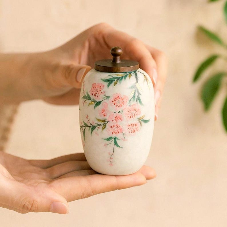 May include: A small, off-white ceramic jar with a bronze-colored lid. The jar is adorned with hand-painted pink floral designs and green leaves. The jar is held in open hands.