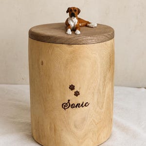 Puede incluir: Una urna cil&iacute;ndrica de madera clara con una tapa de madera oscura coronada por una peque&ntilde;a figura realista de un perro marr&oacute;n y blanco. El frente de la urna tiene grabadas dos peque&ntilde;as huellas de patas sobre el nombre Sonic en letra cursiva.