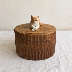 May include: A round wooden box with a dark brown finish and a vertically carved, textured exterior. The lid features a hand-painted, sculpted orange and white cat figurine resting on top, accompanied by a small wooden plaque engraved with Captain Sara.