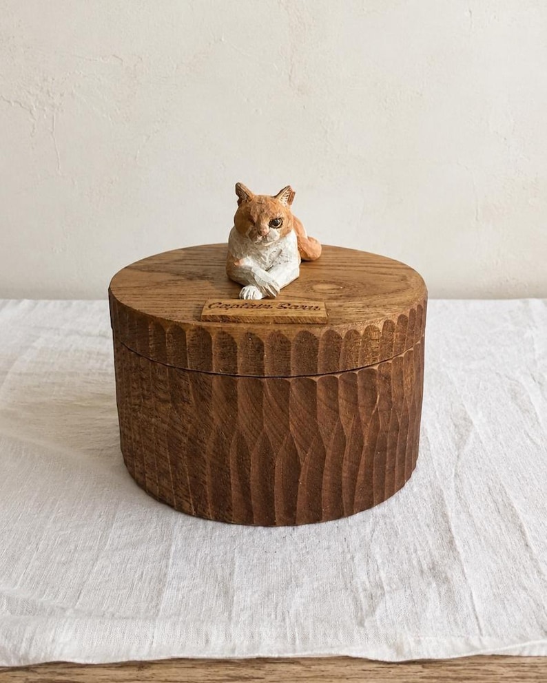May include: A round wooden box with a dark brown finish and a vertically carved, textured exterior. The lid features a hand-painted, sculpted orange and white cat figurine resting on top, accompanied by a small wooden plaque engraved with Captain Sara.