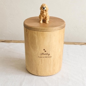 Puede incluir: Una urna cil&iacute;ndrica de madera clara con una tapa de madera oscura rematada por una peque&ntilde;a figura de perro color canela. El frente de la urna est&aacute; grabado con dos huellas, el nombre "Hurley" y el texto "Forever our little Herman" en marr&oacute;n.