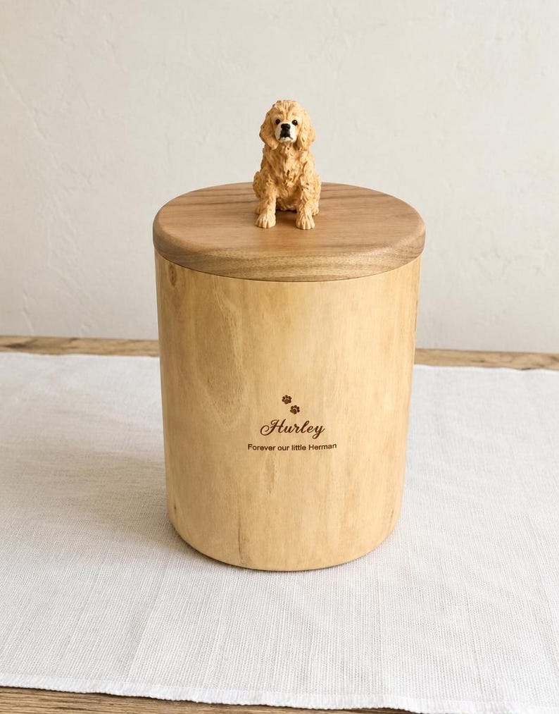 Puede incluir: Una urna cil&iacute;ndrica de madera clara con una tapa de madera oscura rematada por una peque&ntilde;a figura de perro color canela. El frente de la urna est&aacute; grabado con dos huellas, el nombre "Hurley" y el texto "Forever our little Herman" en marr&oacute;n.