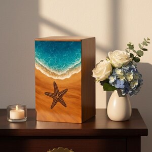 Pode incluir: Urna de madeira com um design de cena de praia. A parte superior apresenta um oceano azul com ondas brancas, que se transforma numa praia arenosa com uma estrela do mar. Uma vela acesa e um vaso de flores estão numa mesa de madeira escura.