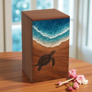 May include: A wooden box with a sea turtle design. The top and sides are a dark wood grain. The front features a beach scene with blue waves and a brown sea turtle. Pink flowers are in the foreground.
