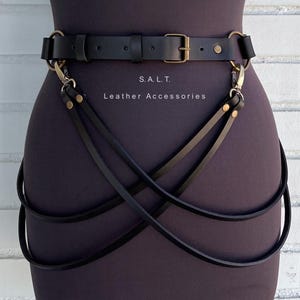 May include: Black leather harness with gold-tone metal hardware, including a buckle and clasps. The harness has straps that cross the body. The text "S.A.L.T. Leather Accessories" is visible.