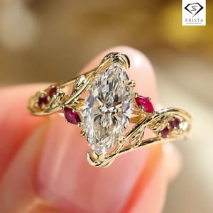 May include: A gold ring featuring a large, oval-cut diamond as the centerpiece, accented with small red gemstones and a delicate, leaf-like design. The ring is set against a soft, blurred background.