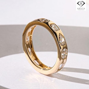 Puede incluir: Un anillo de oro con una serie de diamantes de varias formas. El anillo presenta una mezcla de diamantes redondos, esmeralda y en forma de pera engastados en una banda de oro pulido. El anillo se muestra sobre una superficie blanca.