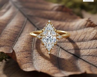 May include: A marquise-cut diamond ring with a gold band and a halo of smaller diamonds. The ring is set on a brown leaf, with the Arista Design Studio logo in the upper right corner.